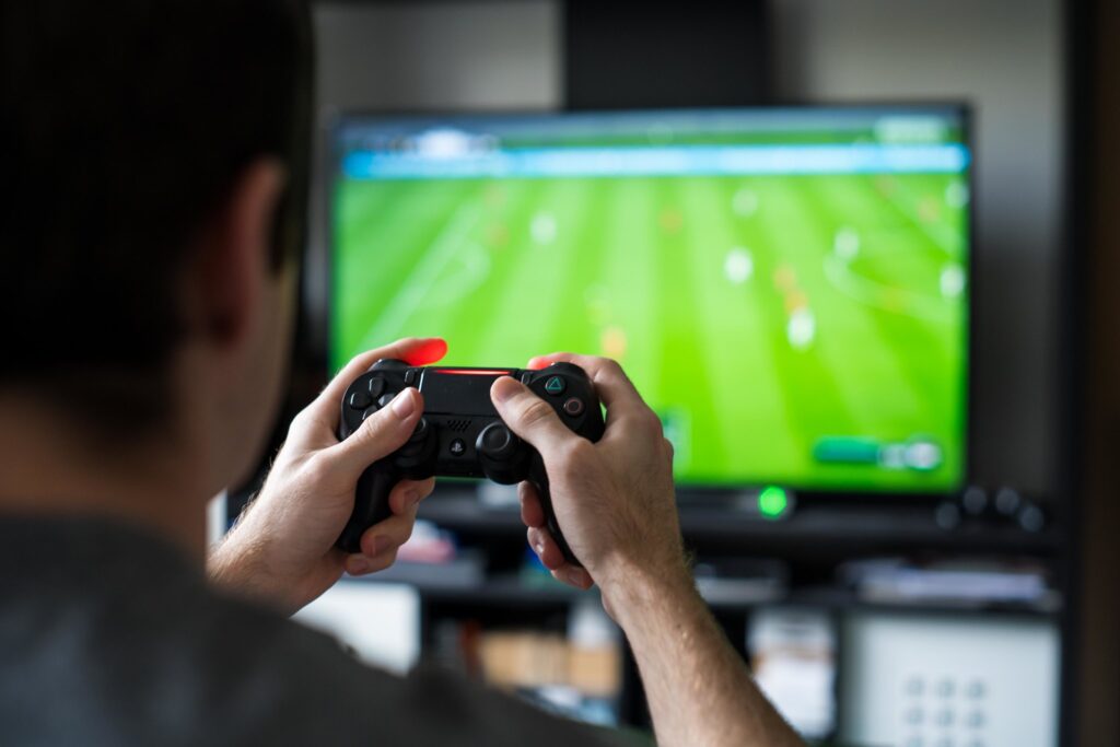 A man is playing videogames, photographed from behind. Only the man's hands and the game controller are in focus. The screen is visible in the background, blurred, but appearing to show a football game.