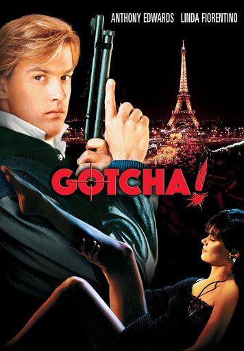 Cover art for the 1985 film "Gotcha!" Anthony Edwards stands on the left side of the image wearing a suit and holding up a pistol. Lina Fiorentino is at the bottom of the image, suggestively lifting her bare leg. Behind them is the Eiffel Tower in lights. The word "Gotcha!" is printed in red text, with the O stylized as a pair of crosshairs.