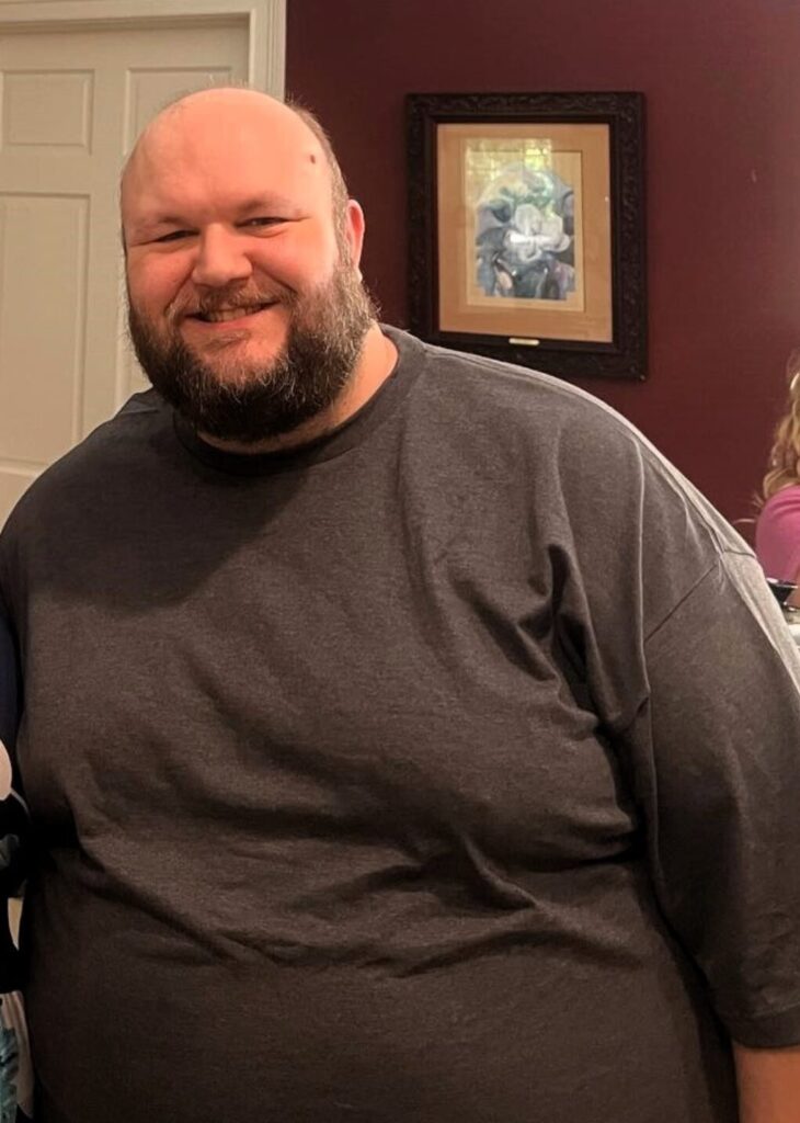 A photo of an overweight man in his late 30s with a bald head and beard, wearing a grey shirt.